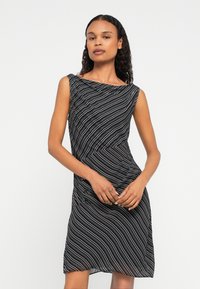 Göndör hajú nő áll egy sima fehér háttér előtt, ujjatlan fekete ruhát visel, amelyen fehér és kék átlós csíkok láthatók.