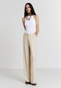 Bodysuit blanc sans manches associé à un pantalon beige à jambes larges. Le pantalon présente une taille à cordon et une texture lisse. Chaussures à plateforme marron.