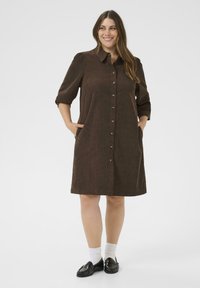 Woman smiling, wearing a brown button-up dress with pockets, white socks, and black loafers, standing against a plain white background.