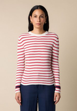 Kobieta o długich ciemnych włosach, nosząca złote kolczyki koła, biały sweter z długim rękawem w czerwone paski oraz granatowe spodnie, stojąca na tle beżowego tła.