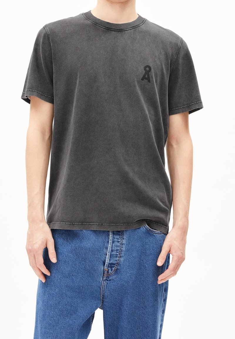 Personne portant un t-shirt noir délavé à col rond avec un petit logo noir sur la poitrine, associé à un jean en denim bleu large.