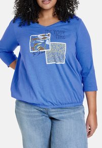 Top in cotone blu con scollo a V e maniche a tre quarti. Presenta due stampe grafiche quadrate e un orlo elastico. Texture liscia.