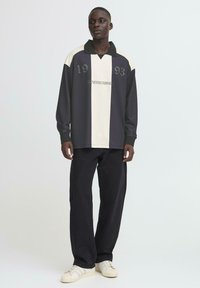 Sort og creme langærmet polo skjorte med krave, kontrasterende paneler og teksten "FREMGANG MÅ FORSIGTIGES" med årene "1993" og "1993."