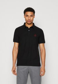 Schwarzes Polohemd mit einem Knopfverschluss mit zwei Knöpfen und kurzen Ärmeln. Mit einem roten gestickten Logo auf der Brust und einem geraden Saum.