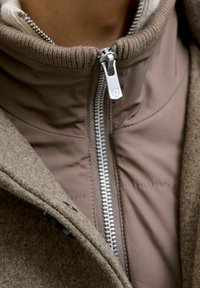 Pruun jakk, millel on pehme, tekstureeritud villane välimus ja sile, polsterdatud sisekiht. Omab hõbedast tõmblukk ja soonikkoes krae.