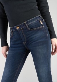 Jeans de mezclilla oscuros de corte ajustado con una textura suave, cierre de botón dorado y costuras destacadas. Detalle de bolsillo con etiqueta de la marca visible.