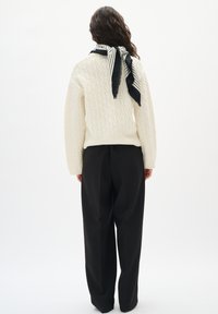 Person med langt krøllet hår iført en cremefarvet cable-knit sweater, sorte brede bukser og et sort-hvidt stribet tørklæde bundet om halsen.