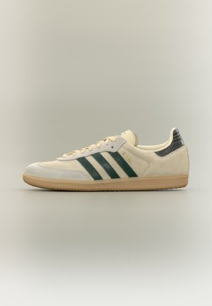 Sneaker in camoscio beige con strisce verde scuro, suola in gomma e linguetta del tallone nera con texture, mostrata di lato su uno sfondo neutro.