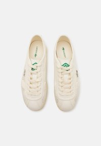 Cream-colored lace-up sneakers made from textured fabric, featuring a green logo, simple shape, and rubber sole. No visible patterns.