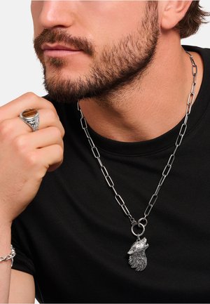 Bärtiger Mann trägt eine silberne Halskette mit einem heulenden Wolf-Anhänger und einen silbernen Ring mit einem braunen Stein an der Hand.