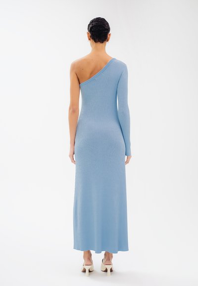 Robe longue bleue côtelée à une épaule, silhouette ajustée, texture douce, longueur mi-mollet, associée à des talons de ton doux.