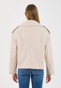 Donna con capelli lunghi e lisci, che indossa un giubbotto di lana beige e jeans blu, mostrata di spalle su uno sfondo bianco semplice.
