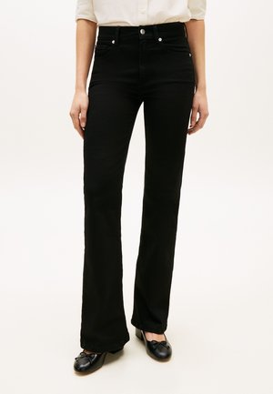 Γυναίκα που φοράει ψηλόμεσα μαύρα τζιν καμπάνα, μαύρα δερμάτινα παπούτσια με χαμηλό τακούνι και φιόγκους, και λευκό πουκάμισο με κουμπιά και μανίκια που έχουν αναδιπλωθεί.