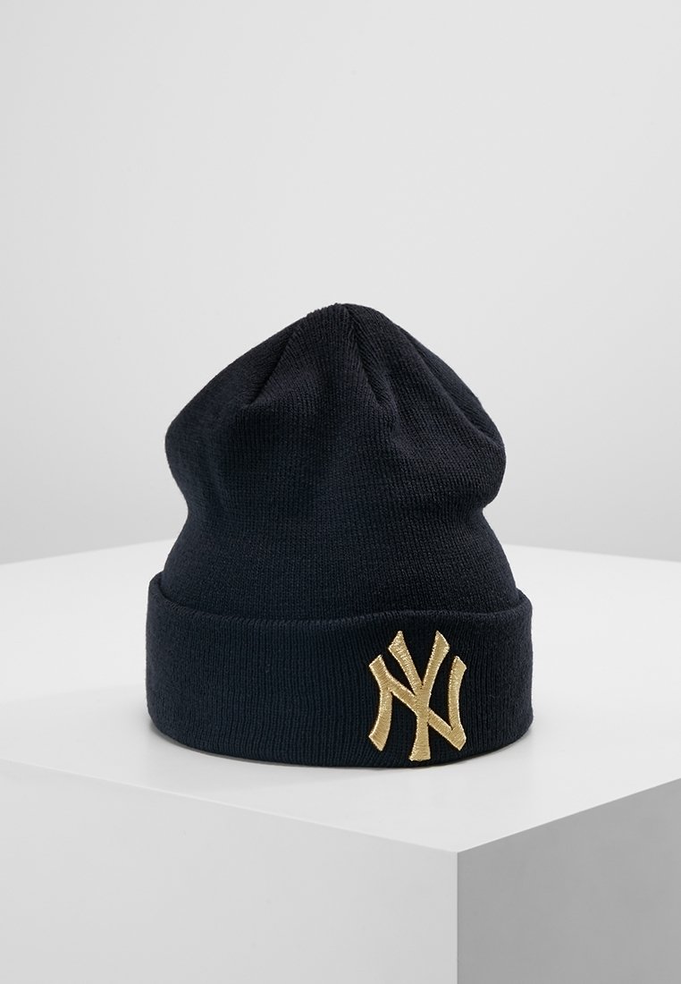 Bonnet en tricot noir avec revers plié arborant le logo des New York Yankees brodé en or, présenté sur un socle blanc sur un fond gris.