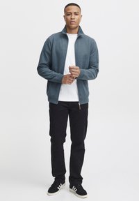 Veste zippée grise avec un col haut, portée sur une chemise blanche, associée à un pantalon noir et des baskets noires. Design simple, sans motifs.
