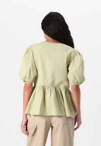 Blusa verde chiaro con maniche a sbuffo, vita arricciata e orlo fluente. Il tessuto sembra morbido e leggero, progettato per un abbigliamento casual.