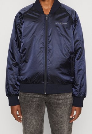 Personne portant une veste bomber zippée Tommy Hilfiger bleu marine avec poignets et ourlet côtelés, associée à un jean gris effet délavé à l'acide.