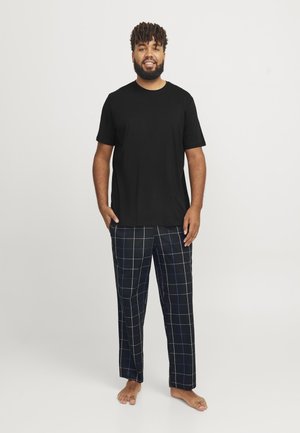 Un tricou negru cu mânecă scurtă asociat cu pantaloni de pijama negri, având un model de grilă albă și albastră, expus pe o persoană care stă desculță.