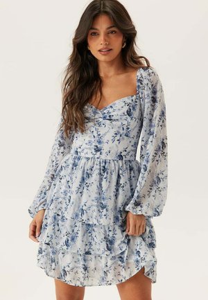 Kvinna klädd i en ljusblå blommig klänning med långa puffärmar och kjol i flera lager, stående mot en enkel bakgrund.