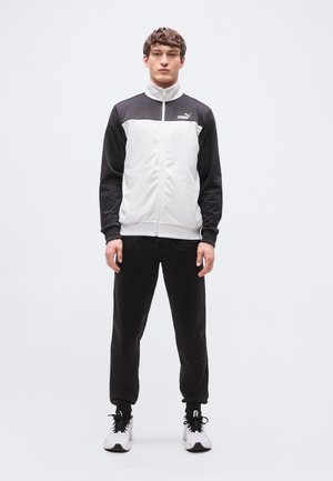 Mladý muž stojaci, oblečený v čierno-bielej športovej bunde Puma, čiernych nohaviciach a bielych teniskách na jednoduchom bielom pozadí.