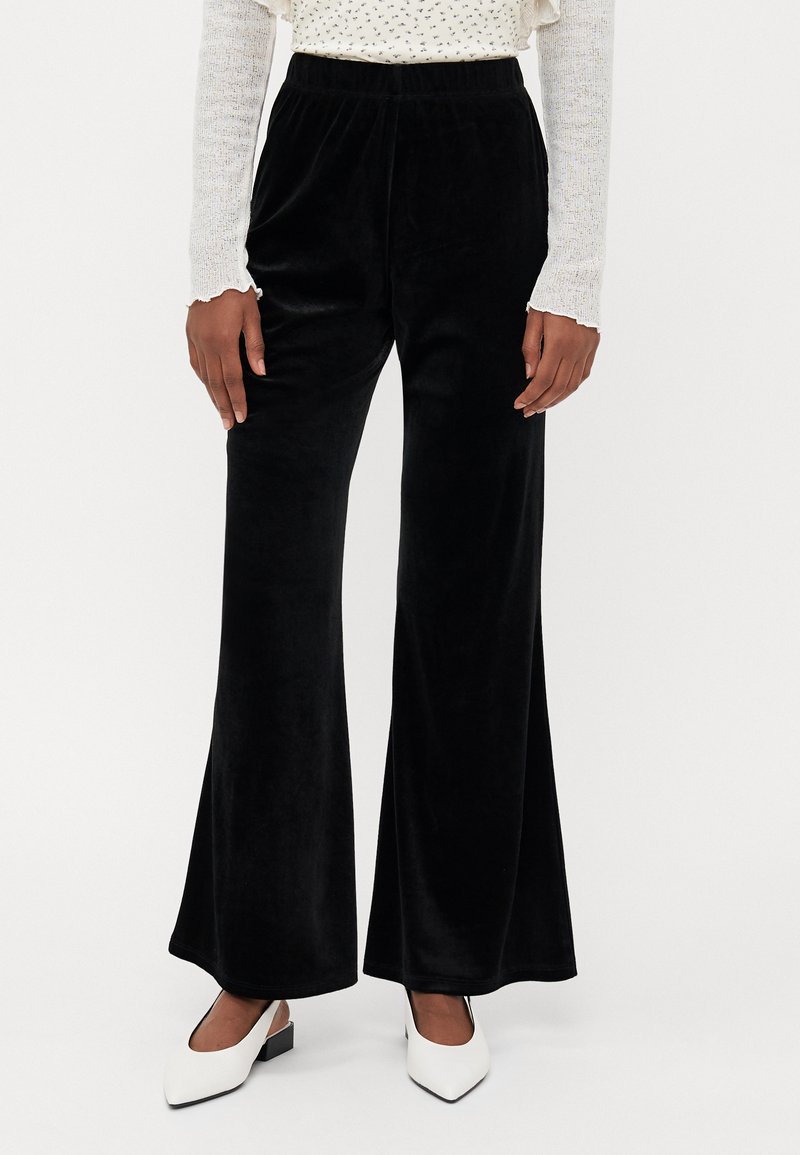 Zwarte, uitlopende velours broek met een elastische tailleband; zachte textuur en volledige lengte; gecombineerd met witte schoenen met een spitze teen.