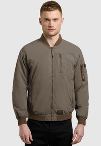 Jeune homme portant une veste bomber marron avec des poches zippées sur la poitrine et les manches, debout devant un fond gris clair uni.