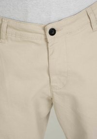 Béžové bavlněné kalhoty s přímým střihem, černým knoflíkem, zesíleným šitím a jednou kapsou na pravé straně.