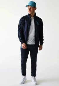 Chándal navy con detalles en azul claro, chaqueta con cremallera y pantalones a juego, combinado con una gorra azul claro y zapatillas blancas.