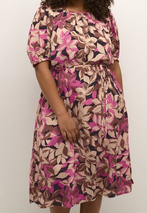Vrouw die een knielange jurk draagt met pofmouwen, met een roze, beige en bruine bloemenprint en een gestrikte tailleband.