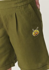 Olivovo zelené šortky z ľahkého materiálu, s bočnou vreckom a žltou nášivkou s karikatúrou postavy a anténami na prednej strane.