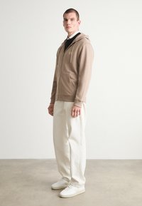Felpa con zip color marrone chiaro, dotata di tasca frontale, abbinata a pantaloni bianchi larghi e sneakers bianche. Branding minimale e design casual.