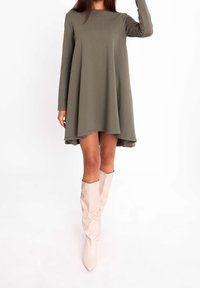 Olivgrünes, langärmliges Kleid mit A-Linien-Silhouette und fließendem Saum. Kombiniert mit kniehohen, hellbeigen Stiefeln mit spitzer Form.