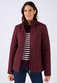 Giacca trapuntata bordeaux con colletto alto, caratterizzata da motivi a rombi e chiusura con zip. Abbinata a un maglione a righe e jeans scuri.