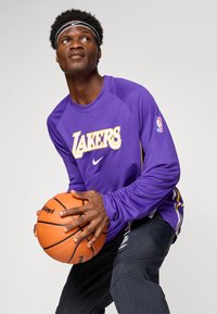 T-shirt de basketball à manches longues violet avec le logo "LAKERS" en jaune, associé à un pantalon de sport foncé. Le modèle tient un ballon de basketball orange et porte un bandeau noir.