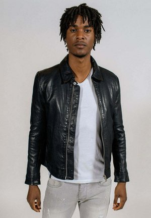 Veste en cuir noire avec col, fermeture éclair à l'avant et coupe ajustée. Portée sur un t-shirt blanc et associée à un jean gris clair.