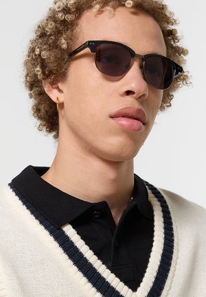 Giovane con capelli biondi ricci che indossa occhiali da sole neri, un orecchino a cerchio dorato, una camicia nera con colletto e un maglione bianco lavorato a maglia con righe blu navy.