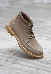 Beige semskede ankelstøvler med rund tå, snørebåndsdesign, gummisåle og kontrastfarget hvit søm. Teksturert overflate med lett glans.
