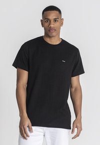 Czarny t-shirt o teksturowanej powierzchni, z krótkim rękawem, żebrowanym wzorem, okrągłym dekoltem oraz małym logo na piersi, noszony z białymi spodniami.