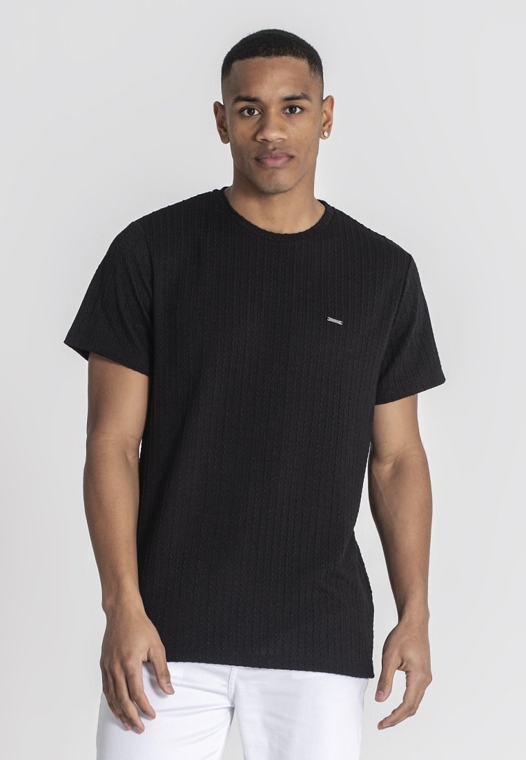 Crna teksturirana majica kratkih rukava, rebrasti uzorak, okrugli izrez, mala oznaka na prsima, uparena s bijelim hlačama.