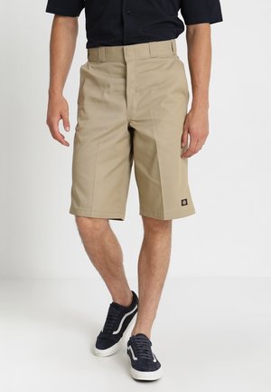 Shorts cargo beiges à coupe droite, devant plat et petite écusson logo sur la jambe gauche. Portés avec une chemise bleu marine et des baskets.