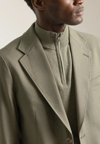 Blazer de color verde oliva claro con solapas de muesca, combinado con un top de punto de cuello alto a juego que presenta un cierre de cremallera. Textura de tela suave.