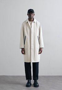 Ljusbeige trenchcoat med krage och sidofickor, kombinerad med svarta byxor och skor. Modellen bär en svart keps. Enkel design.