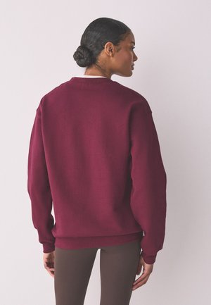 Kobieta z ciemnymi włosami związanymi w kok, ubrana w jednolity bordowy sweter z długim rękawem i ciemne spodnie, stoi tyłem na neutralnym tle.