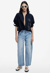 Marineblaues Hemd mit Knöpfen und floralen Stickereien, gerade geschnittene hellblaue Jeans und schwarz-weiße Slipper. Weiße rechteckige Sonnenbrille.