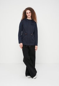 Veste à capuche marine avec deux poches devant et cordons ajustables. Portée avec un pantalon noir à jambes larges et des baskets blanches. Texture lisse.