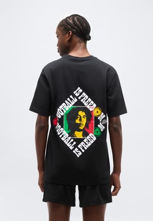 Άτομο με πλεγμένα μαλλιά που φοράει μαύρο σορτς και μαύρο T-shirt με πολύχρωμο γραφικό και το κείμενο «Το ποδόσφαιρο είναι ελευθερία» στην πλάτη.