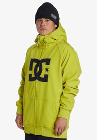 Limegrön huvtröja med dragkedja fram, stor svart logotyp, justerbar huva och ribbade ärmslut. Buren med en svart stickad mössa.