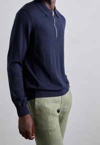 Mörkblå långärmad polotröja med dragkedja vid kragen, tillverkad av mjukt material, ihop med ljusgröna linbyxor.