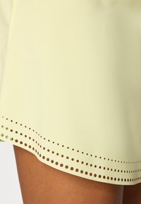 Vestido amarelo claro com um design amplo, apresentando uma bainha adornada com recortes circulares espaçados uniformemente, textura suave.
