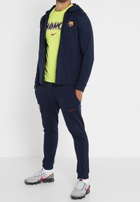 Felpa blu con zip e dettagli rossi, abbinata a una maglietta gialla neon con grafica, pantaloni jogger blu navy e scarpe da ginnastica grigie.